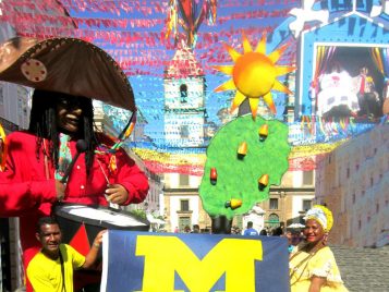 A man and a woman hold either end of a block-M flag. They have put down their baskets to hold it. The woman is wearing a yellow-and-white dress with a large hoop skirt. The man is wearing a yellow shirt and blue shorts. A person dressed in a tall costume with a brown hat and a bright red jacket is playing the drums behind them. They are standing in front of a colorful mural of the sun, the sky, and an old building.